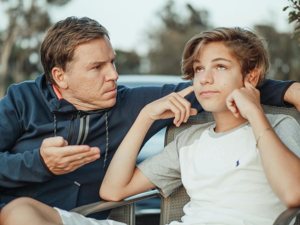 pexels-photo-8550837-8550837 Father expressing frustration while teenage son shows defiance, seated outdoors.